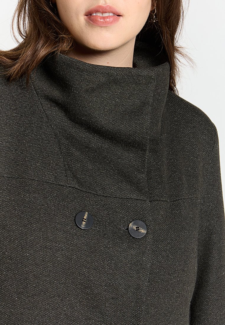 Veste sombre et texturée avec un col montant, deux boutons noirs et un design structuré. La couleur est principalement noire avec des variations tonales subtile.