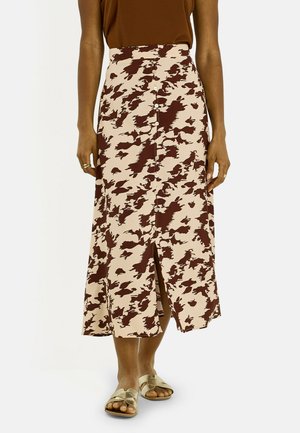Femme portant une jupe midi beige et marron à motifs avec des boutons sur le devant et une fente à l'avant, associée à des sandales slides dorées.