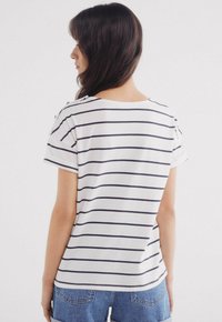 White t-shirt with navy horizontal stripes, short sleeves, rounded neckline, and button accents on shoulder. Paired with denim shorts.