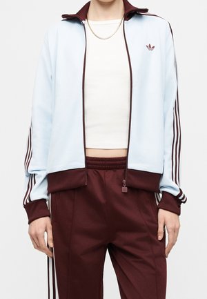Person iført lyseblå og mørkerød Adidas træningsjakke med matchende mørkerøde træningsbukser og en hvid kort top, synlig fra nakke til midt på låret.