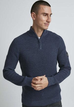 Marineblå ribbestrikket genser med kvart-glidelås, tettsittende mansjetter og en teksturert design. Har et lite logopatch på overarmen.