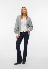 Cárdigan de punto gris claro con mangas anchas, llevado sobre una camisa blanca de botones y jeans acampanados de color azul oscuro, con un fondo liso.
