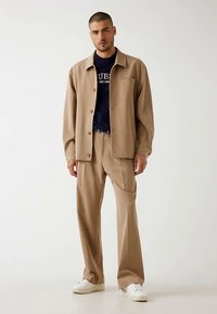 Beige Jacke mit Knopfleiste und Brusttasche, kombiniert mit beigen Hosen, mit klarem Design und weißen Turnschuhen. Darunter ein einfarbiges marineblaues Hemd.