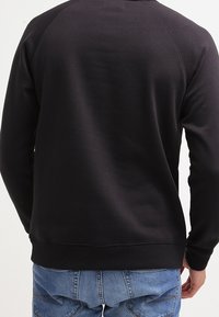 Svart crewneck-tröja med ribbade ärmslut och nederkant, med en slät yta och raglanärmar, bärs över blå jeans.