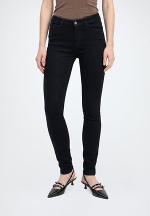 Femme portant un jean skinny noir, un haut marron et des chaussures noires pointues à talon bas avec des brides à la cheville, debout devant un fond uni.