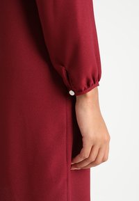 Robe bordeaux en tissu lisse et texturé ; possède une manche ample avec un accent de bouton près du poignet et de la couture latérale.