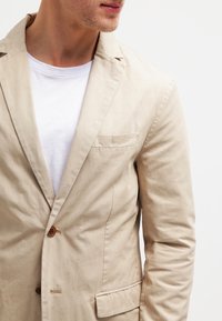 Blazer di cotone beige con una tasca sul petto, frontale a due bottoni e una texture liscia, indossato sopra una T-shirt bianca.