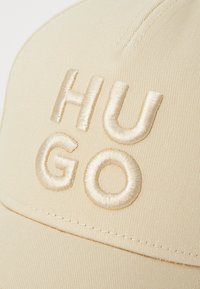 Beige baseball cap featuring raised, embroidered letters "HUGO" in a matching satin finish. The fabric is textured, with visible stitching.