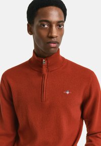 Röd zip-up tröja gjord av textil med struktur, med hög krage och en liten logo på bröstet. Slät yta, figurnära design.