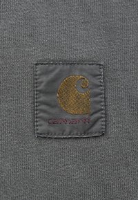 Γκρίζος ύφασμα που διαθέτει ένα τετράγωνο ύφασμα με κίτρινο κεντημένο λογότυπο και κόκκινο κείμενο που αναγράφει "Carhartt." Ορατές ραφές γύρω από το ύφασμα.