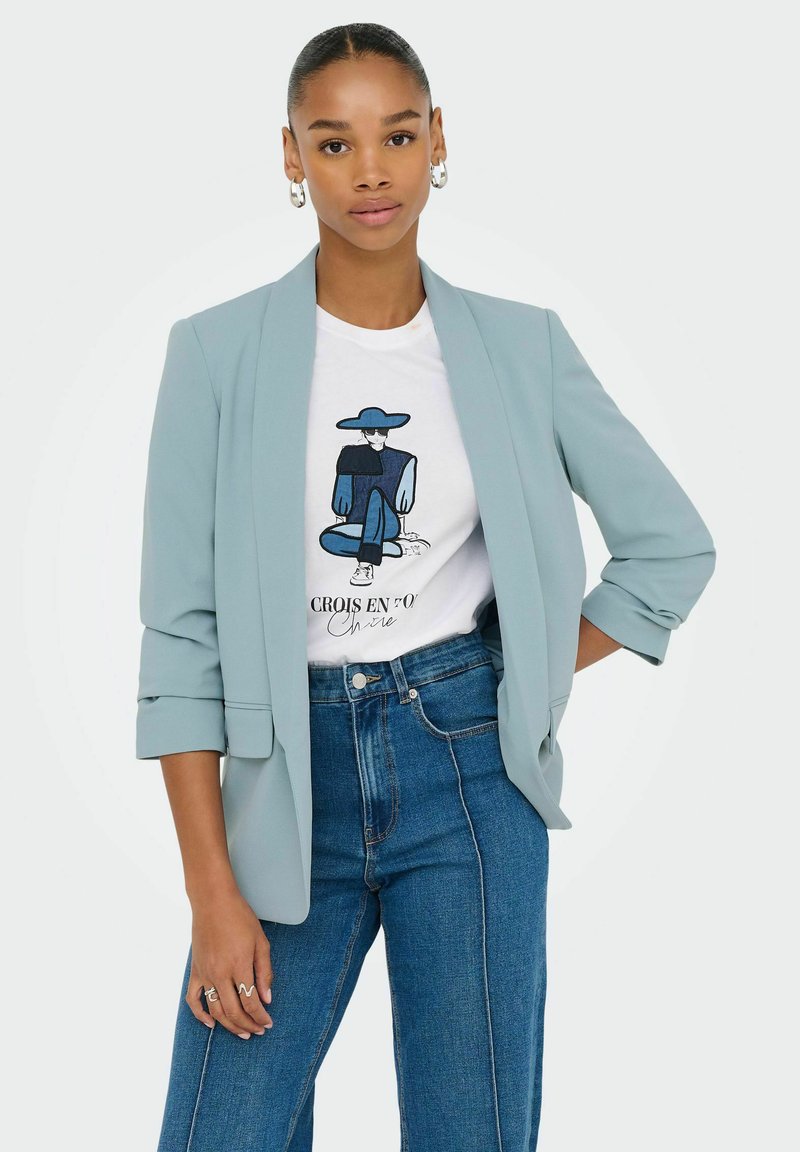 Blazer azzurro chiaro con maniche arrotolate, indossato sopra una t-shirt bianca con un design grafico blu, abbinato a jeans in denim a vita alta.