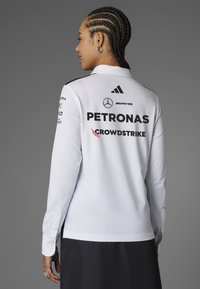 Vit långärmad polo med svarta accenter, prydd med logotyper på ryggen: Mercedes, Petronas och Crowdstrike. Knäppta ärmslut.