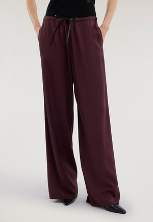 Kvinde iført løstsiddende burgunderrøde bukser med sort snor, hænder i lommerne, sorte spidse sko og sort top, stående på neutral baggrund.