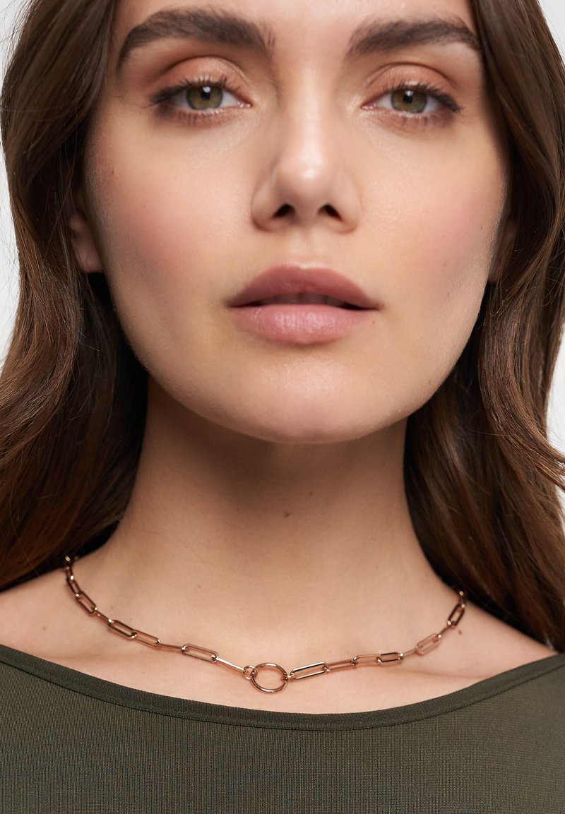 Halsband av roséguld med avlånga länkar och en cirkulär detalj i mitten, bärs runt halsen mot en mörkgrön topp.