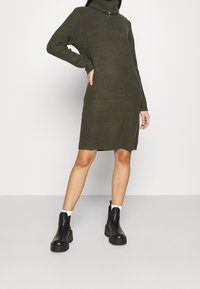 Olivenfarvet ribstrikket kjole med høj funnel neck og halv lynlås lukning. Modellen parrer den med sorte ankelstøvler.