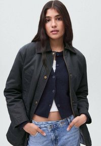 Ung kvinna med långt mörkt hår som bär en svart jacka över en marinblå cardigan och ljusblå jeans, händerna i fickorna, neutral bakgrund.