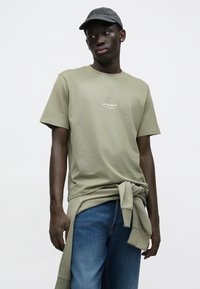 Oliwkowa zielona bawełniana koszulka z krótkim rękawem i logo z przodu, zestawiona z niebieskimi dżinsami i ciemną czapką, z detalem w postaci zawiązanego sweatshirtu.