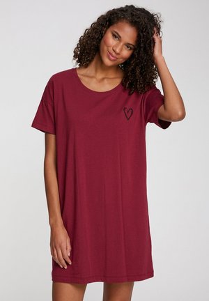 Frau mit lockigem Haar, die ein lockeres bordeauxfarbenes T-Shirt-Kleid mit einem kleinen schwarzen Herz auf der Brust trägt, lächelt und berührt ihr Haar.
