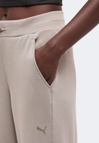 Gros plan d'une personne portant un pantalon de jogging beige Puma avec une main dans la poche latérale, montrant la taille et une partie d'un haut noir.