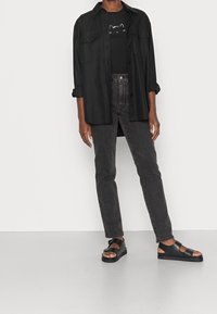 Camisa negra de botones sobre una camiseta gráfica de un gato negro, combinada con jeans oscuros y sandalias negras. Tela suave y diseño sencillo.