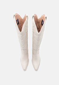Bottes de cowboy en cuir blanc avec des orteils pointus, des motifs de couture complexes, et une tige haute avec un bord découpé. Intérieur doublé de beige clair.