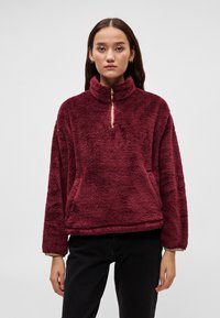 Bordowy polarowy pulower z wysokim kołnierzem, półzamkiem i miękkim, teksturowanym materiałem. Przednia kieszeń typu kangurek i luźne rękawy.