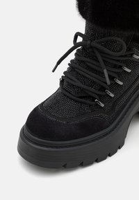 Botte d'hiver noire avec semelle épaisse, bout rond, œillets en métal, lacets noirs, tissu texturé et bordure en fourrure en haut.