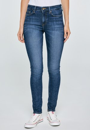 Žena v tmavě modrých úzkých džínách a bílých nízkých teniskách s červenými a černými detaily, stojící na světlém pozadí.