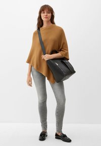 Poncho a maglia color ruggine con vestibilità ampia, abbinato a jeans slim-fit grigi. Borsa in pelle nera con cerniera e manici corti. Mocassini neri.