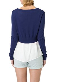 Námořnická modrá pletená halenka s dlouhým rukávem a zkráceným střihem, navrstvená přes bílou blůzu, nošená s světle zelenými šortkami. Hladká textura, přiléhavý design.