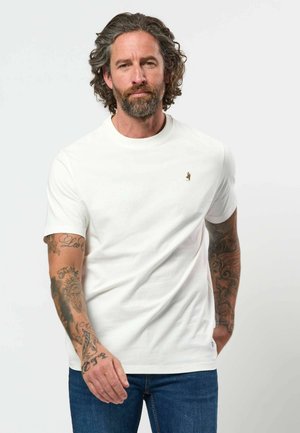 Man met krullend haar en baard, die een wit T-shirt en blauwe spijkerbroek draagt, met getatoeëerde armen, staand voor een eenvoudige achtergrond.