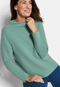Mintgroene textuur top met een hoge hals, lange mouwen en geribbelde uitstraling. Gecombineerd met donkerblauwe jeans in een casual pose.