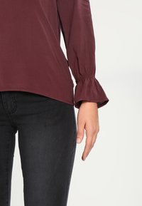Blusa marrone con maniche a campana, tessuto morbido e orlo asimmetrico abbinata a jeans in denim scuro. Focus sulla texture e sul contrasto di colore.