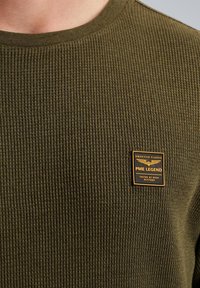 Pull côtelé vert olive avec un col rond, arborant un patch logo PME Legend noir et doré sur le devant. Tissu texturé.