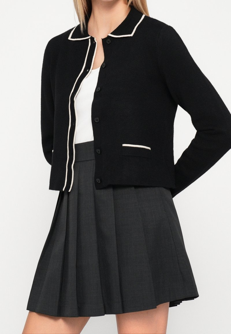 Femme portant un cardigan noir avec un bord blanc, un haut blanc et une jupe plissée gris foncé, debout les mains derrière le dos.