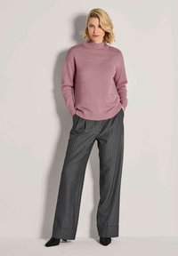 Rosa Strickjacke mit hohem Kragen und gerippten Akzenten, kombiniert mit dunkelgrauen, gestreiftenweiten Hosen und schwarzen, spitzen Schuhen.