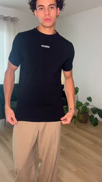 Fiatal férfi göndör hajjal, fekete Karl Lagerfeld pólót és bézs nadrágot visel, beltéren, egy cserepes növény és egy kanapé közelében áll.