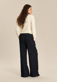 Witte ribgebreide trui en donkerblauwe wijde jeans. De jeans hebben een elastische tailleband, wat een ontspannen silhouet benadrukt.