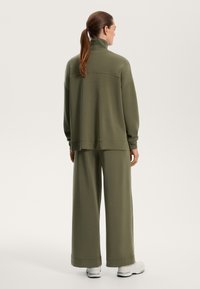 Olivgrön tröja med hög krage och vida ärmar, i kombination med matchande vida byxor, båda i mjukt material.
