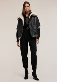 Kobieta w czarnych spodni wysokiego stanu, czarnych botkach na obcasie, ciemnobrązowej koszuli na guziki oraz czarnej skórzanej kurtce z białym kołnierzem z owczej wełny.