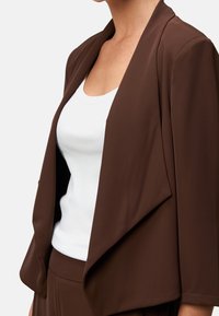 Ruskea bleiseri, jossa on huivikaulus, vino leikkaus ja räätälöity istuvuus, kerrospukeutuneena valkoisen toppipaidan päälle. Sileä kangaspinta, moderni muotoilu.
