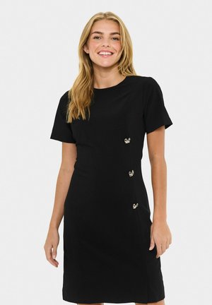 Donna sorridente con lunghi capelli biondi che indossa un vestito nero a maniche corte con tre bottoni argentati a forma di fiocco sul lato.