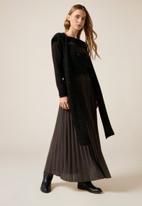 Svart långärmad topp med ett glittrande stickat överlägg, kombinerad med en grå plisserad maxikjol och svarta ankelstövlar, stående på en ljus bakgrund.