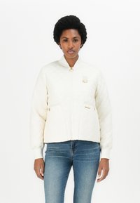 Gepolsterte cremefarbene Bomberjacke mit Reißverschluss, Druckknopftaschen, gerippten Bündchen und einem goldenen Hardware-Akzent am Kragen. Kombiniert mit Jeans.