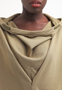 Prenda de tela drapeada en verde oliva con textura suave, que presenta un escote drapeado y detalles en metal. Líneas limpias y diseño en capas.