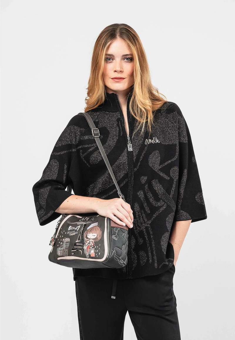 Sudadera negra con cremallera y un patrón texturizado; lleva una bolsa gris claro con un diseño de caricatura y acentos. Estilo de transporte sobre el hombro del modelo.