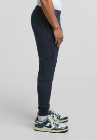 Námornícke jogger shorts s úzkym strihom, elastickým pásom, bočnou vreckou na zips a textúrovanou tkaninou. Skombinované s bielymi a zelenými teniskami.