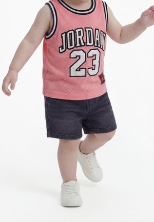 Małe dziecko ubrane w różowy bezrękawnik sportowy Jordan 23, ciemne spodenki i białe trampki, stoi z jedną stopą uniesioną nad ziemią.