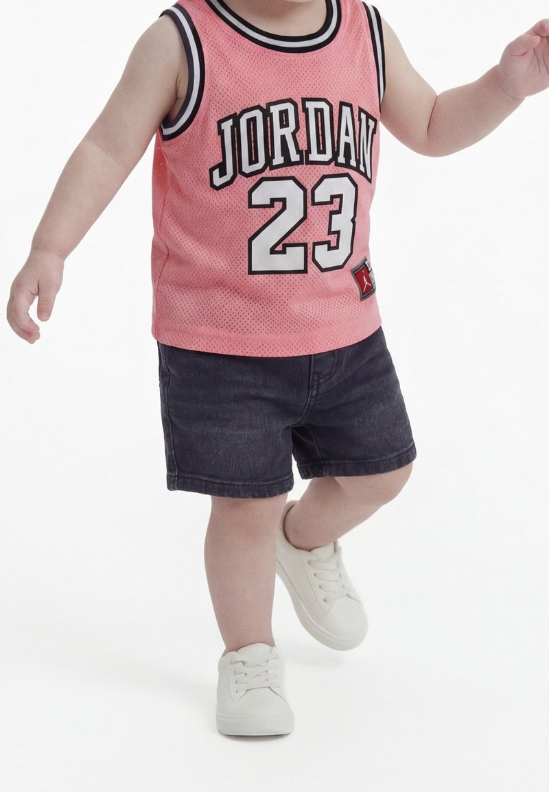 Tout-petit portant un maillot de sport sans manches rose Jordan 23, un short foncé et des baskets blanches, debout avec un pied levé du sol.