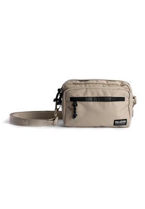 Sac bandoulière rectangulaire beige avec poche zippée sur le devant, sangle ajustable et texte "Dare to Discover" au-dessus de la poche. Étiquette de la marque "Palladium" sur le devant.